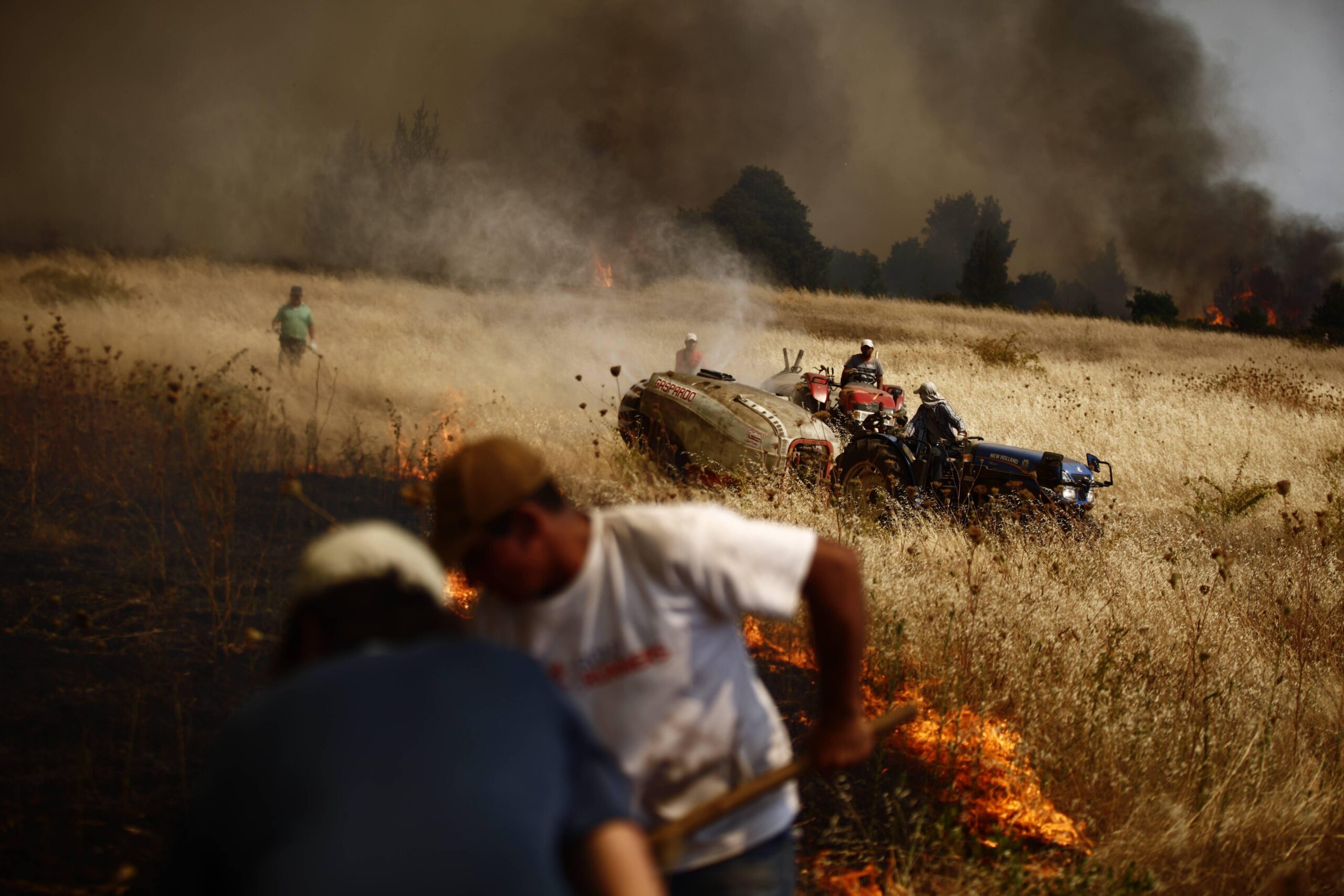 People walk through wildfires in Chile. There is smoke and flames over a wheat field. The sky is black and brown with thick smoke. 