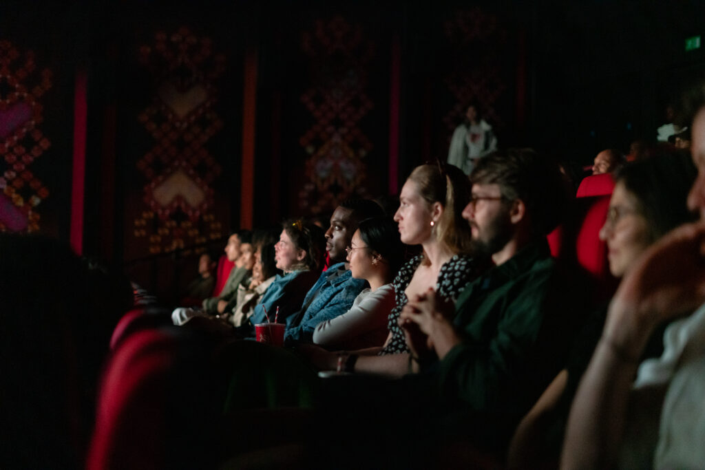 The audience in the cinema sit in the dark on red velvet chairs watching the film. 